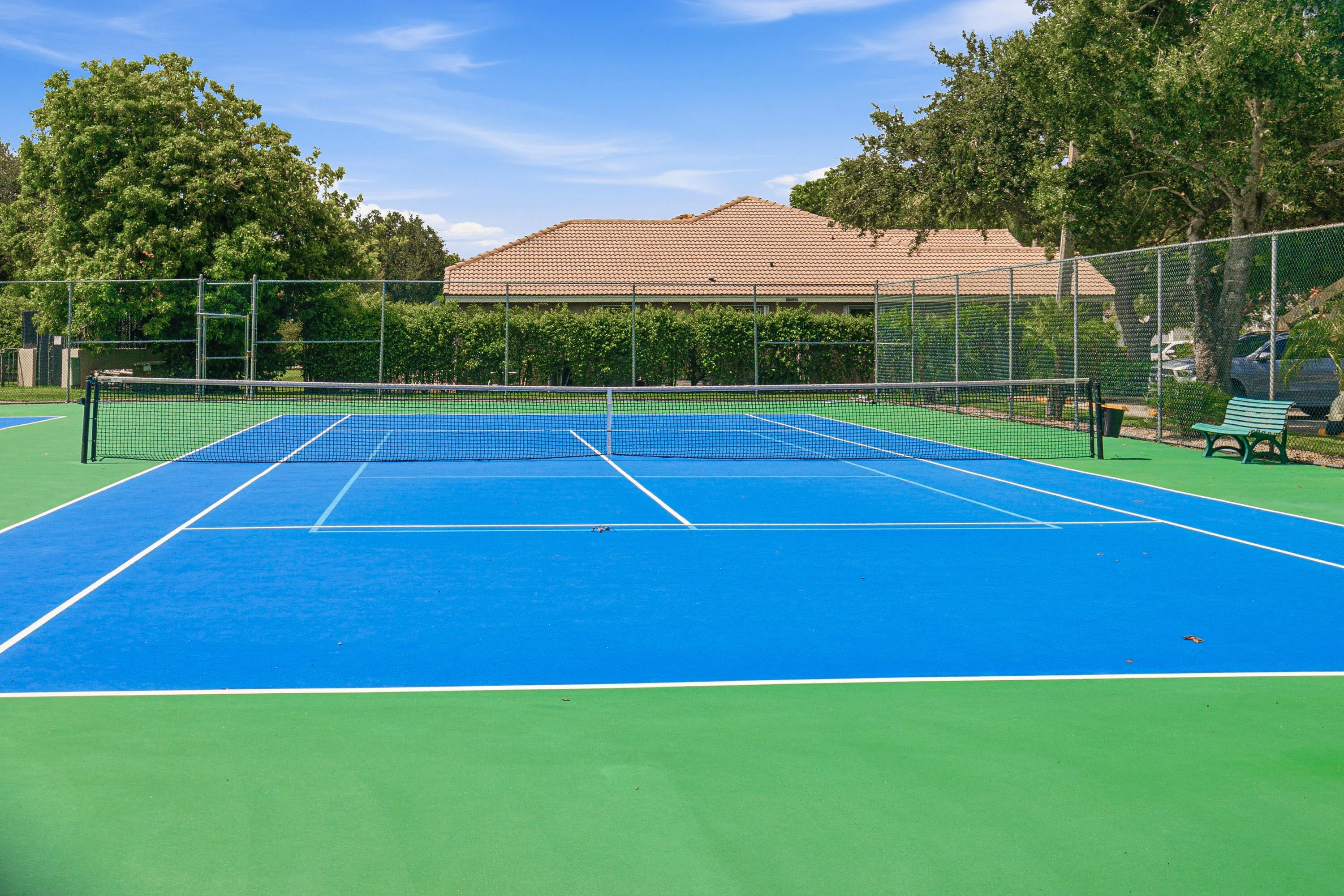 Tennis court featuring a blue playing surface with white lines and green perimeter, surrounded by a chain-link fence