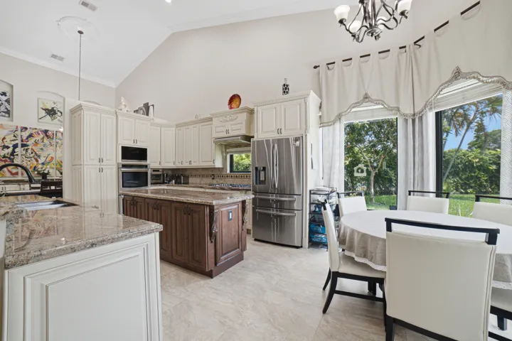 Vaulted ceiling kitchen featuring cream-colored cabinetry, a dark wood-finish island with a stone countertop, integrated stainless steel appliances, and large windows overlooking exterior greenery