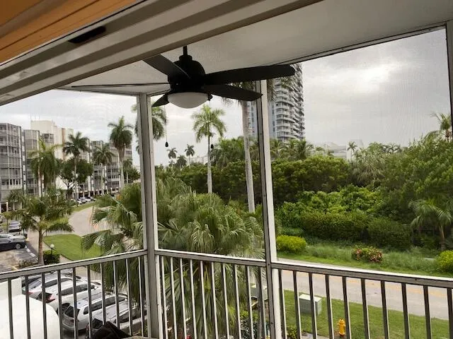 Screened balcony featuring a ceiling fan with integrated lighting, white railing, and exterior views of tropical foliage and distant residential buildings