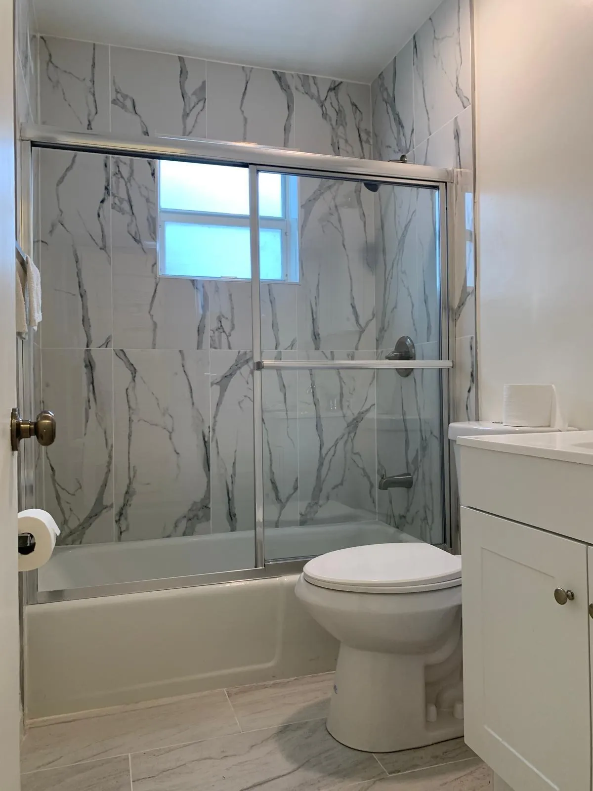 Bathroom featuring a shower-tub combination with sliding glass doors, white and grey veined wall tile, a window, a toilet, and a white vanity with cabinet storage