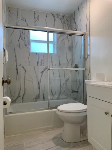 Bathroom featuring a shower-tub combination with sliding glass doors, white and grey veined wall tile, a window, a toilet, and a white vanity with cabinet storage