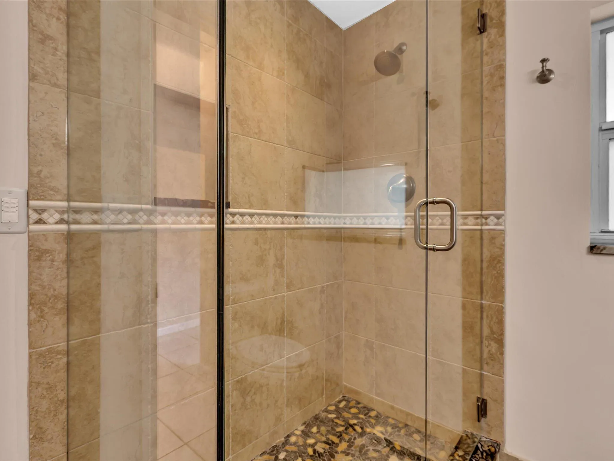 Glass-enclosed shower featuring natural stone-finish wall tile, decorative mosaic tile band, pebble-finish shower floor, recessed shower niche, and brushed chrome fixtures