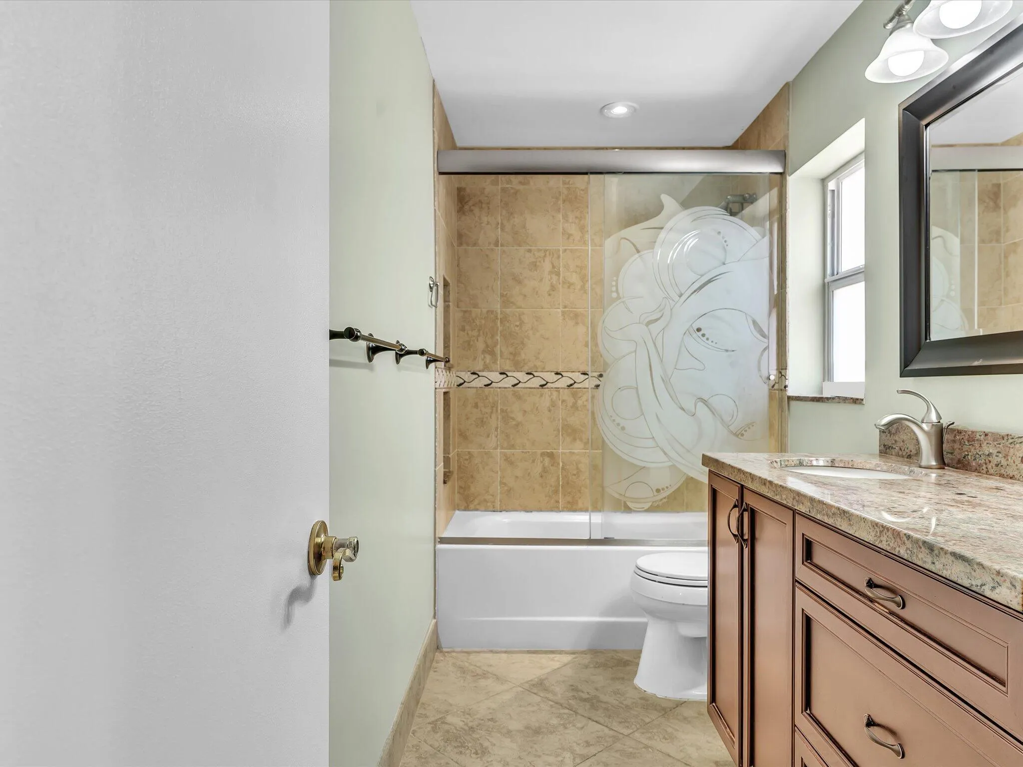 Bathroom featuring a wood-finish vanity with a stone countertop, a glass-enclosed shower/tub with decorative etching, tiled shower walls, and a window for natural light