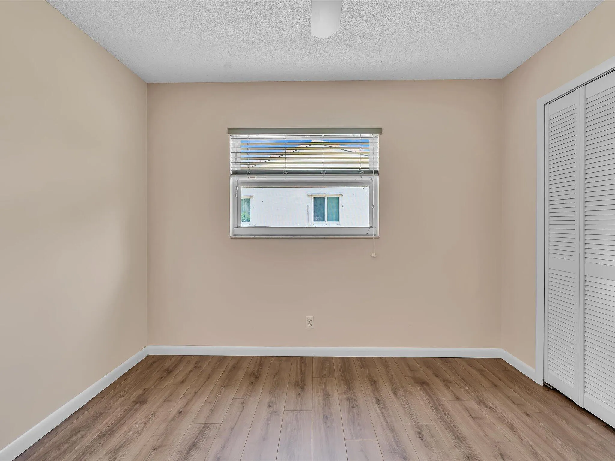 Wood-finish flooring throughout, featuring a single window with blinds, a white ceiling fan, and bi-fold louvered closet doors