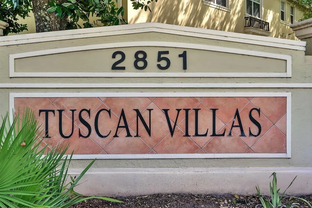 Entry monument featuring tan stucco, a prominent tiled sign with "TUSCAN VILLAS" in black lettering, and dark numerical address "2851."