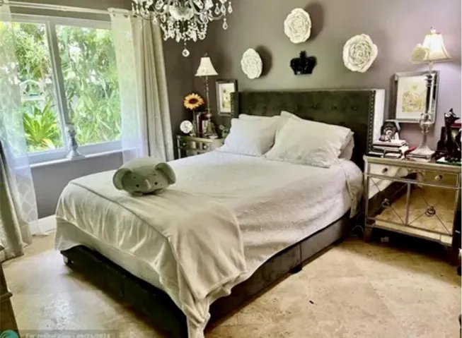 Bedroom featuring a tufted headboard, crystal chandelier, mirrored nightstand, and a large window