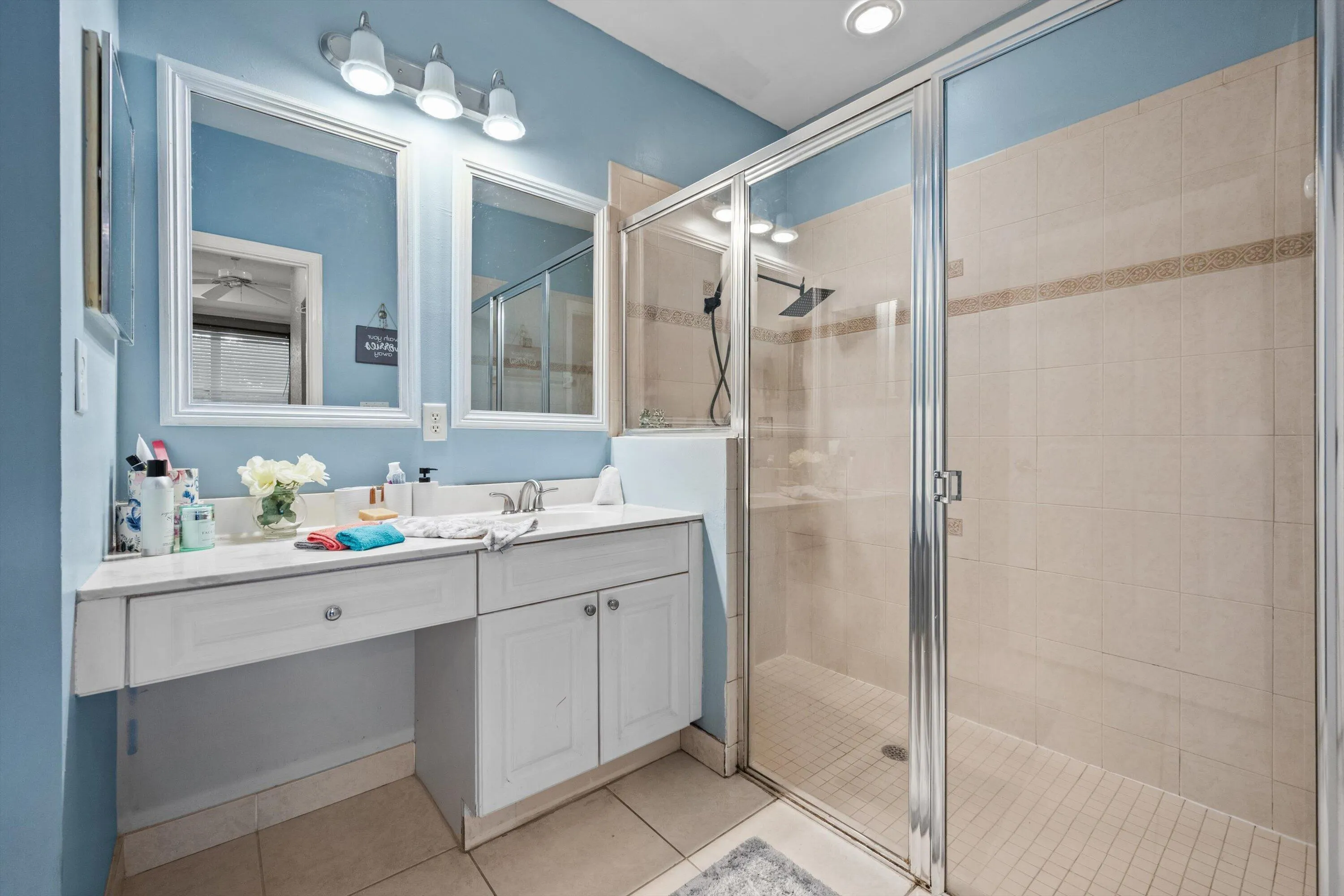 Bathroom featuring a glass-enclosed shower with tile surround, a single vanity with a white countertop and integrated sink, white cabinetry, and two wall-mounted mirrors with vanity lighting