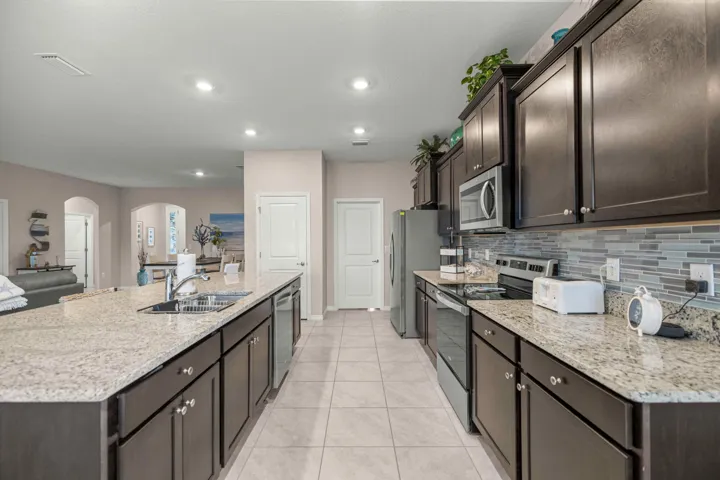 Kitchen featuring an expansive island with a double sink, granite countertops, dark wood cabinetry, a mosaic tile backsplash, and stainless steel appliances including a microwave, range, and refrigerator