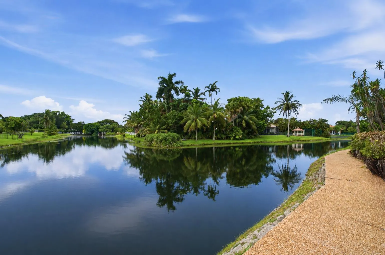 Expansive waterfront setting featuring a tranquil lake, lush tropical landscaping, mature palm trees, and a paved pathway