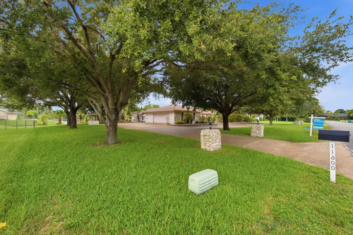 Expansive green lawn with mature trees, a paved driveway, and stone-clad entryway pillars