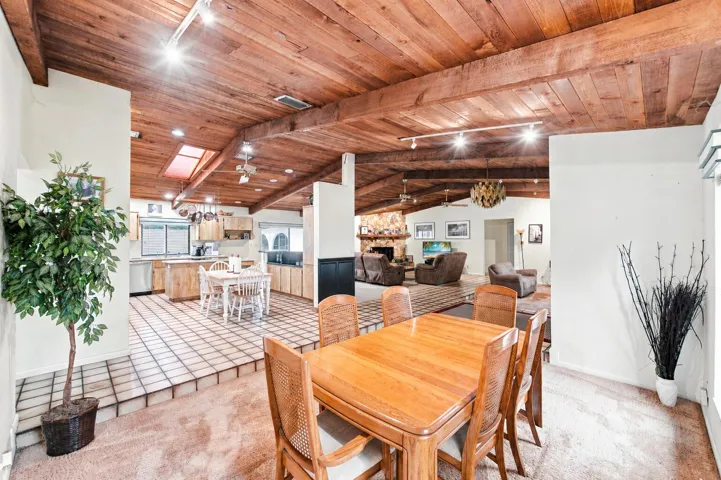 Vaulted wood-paneled ceilings with exposed beams throughout an open-concept interior featuring a kitchen with light wood cabinetry and a skylight, a dining area with wood-finish flooring, and a stone-faced fireplace