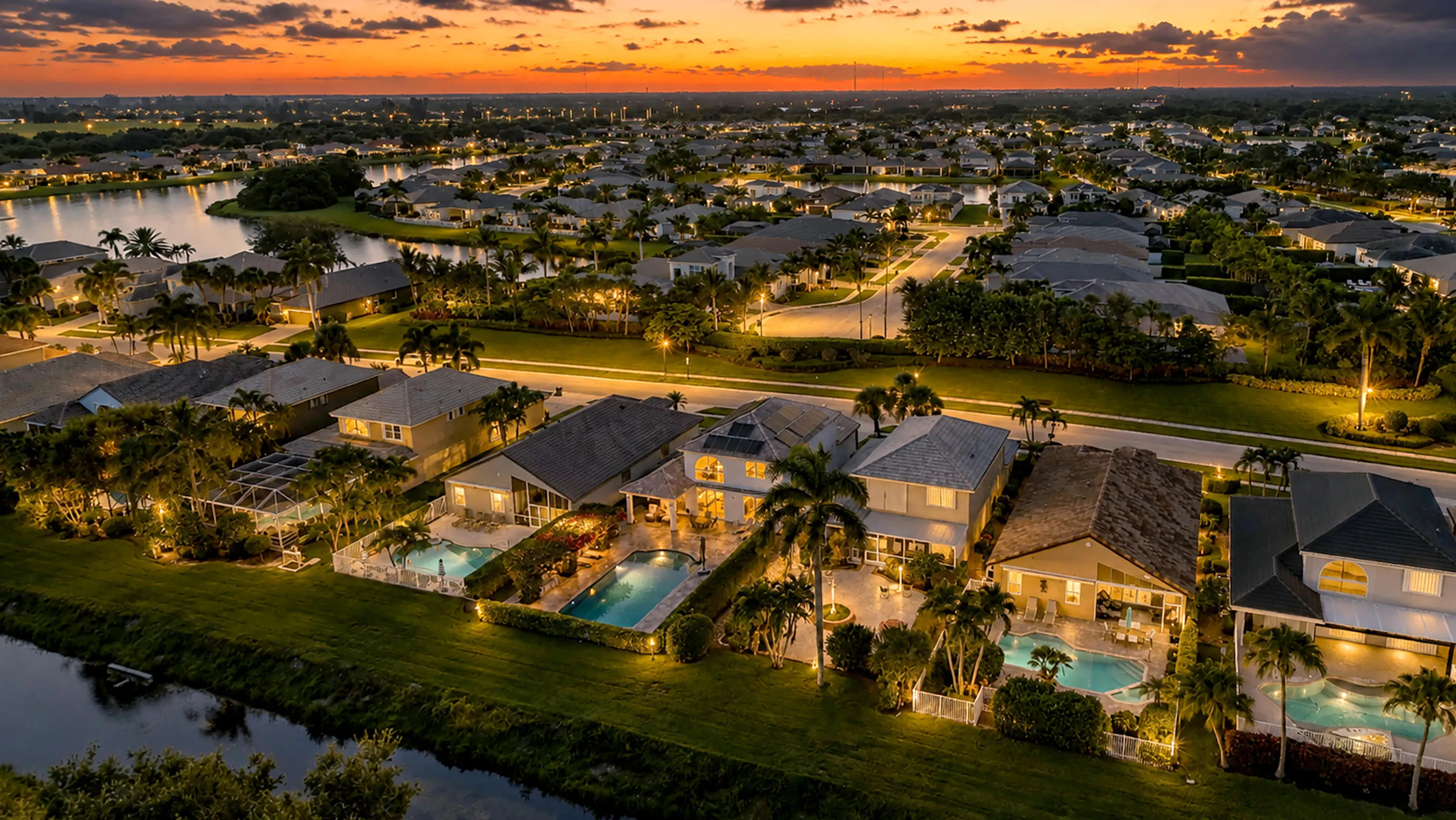 Sunset aerial along the waterway behind the home with illuminated pools and lake views across the community.