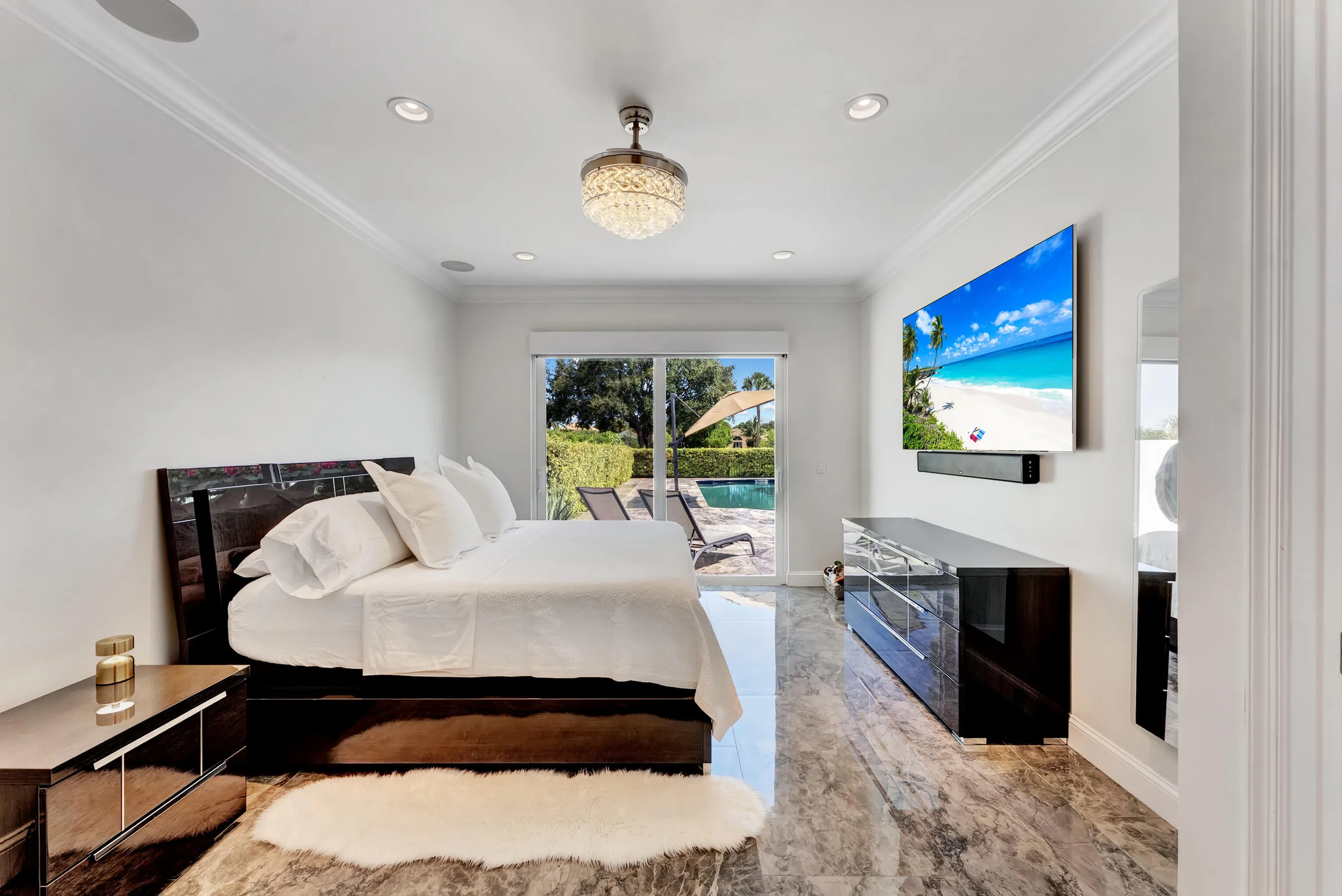 First-floor primary suite with sliding glass doors to the pool deck, mounted TV, and spa-inspired neutral palette.