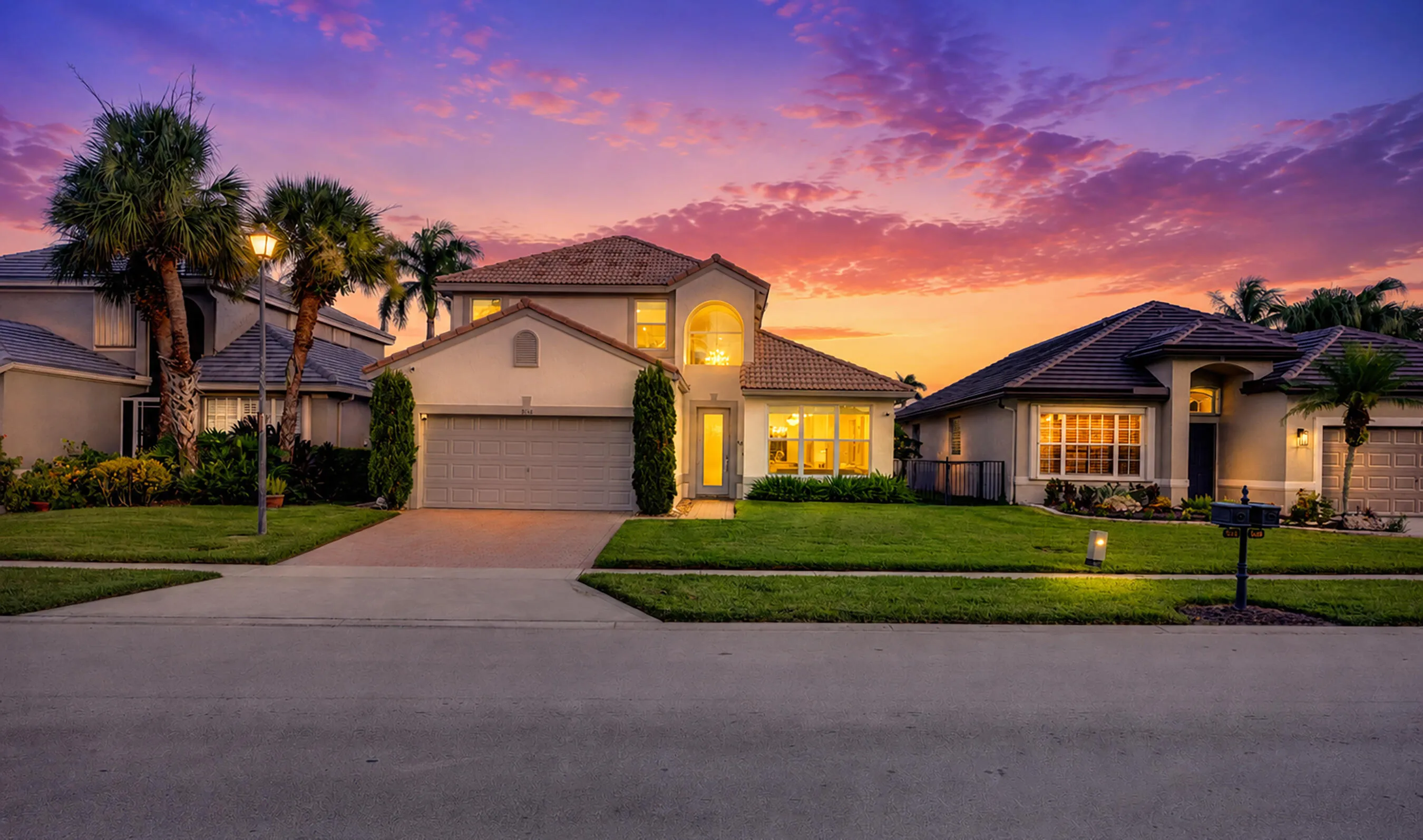 Twilight front exterior of this two-story pool home in Polo Trace, Delray Beach, with paver driveway, lush tropical landscaping, and warm interior lighting at dusk.