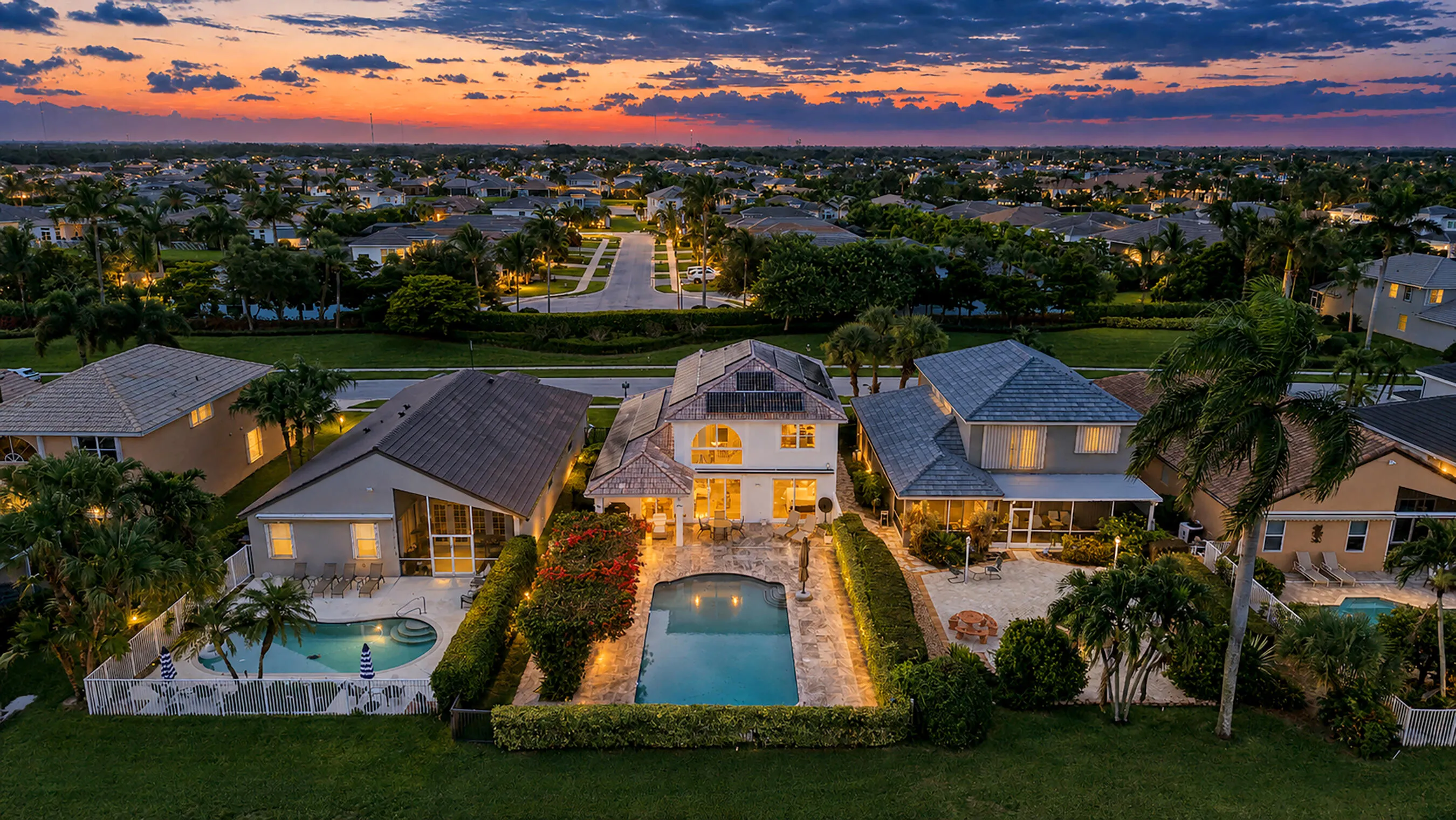 Wide twilight aerial of the home, pool, and neighboring lake community under a fiery sunset.