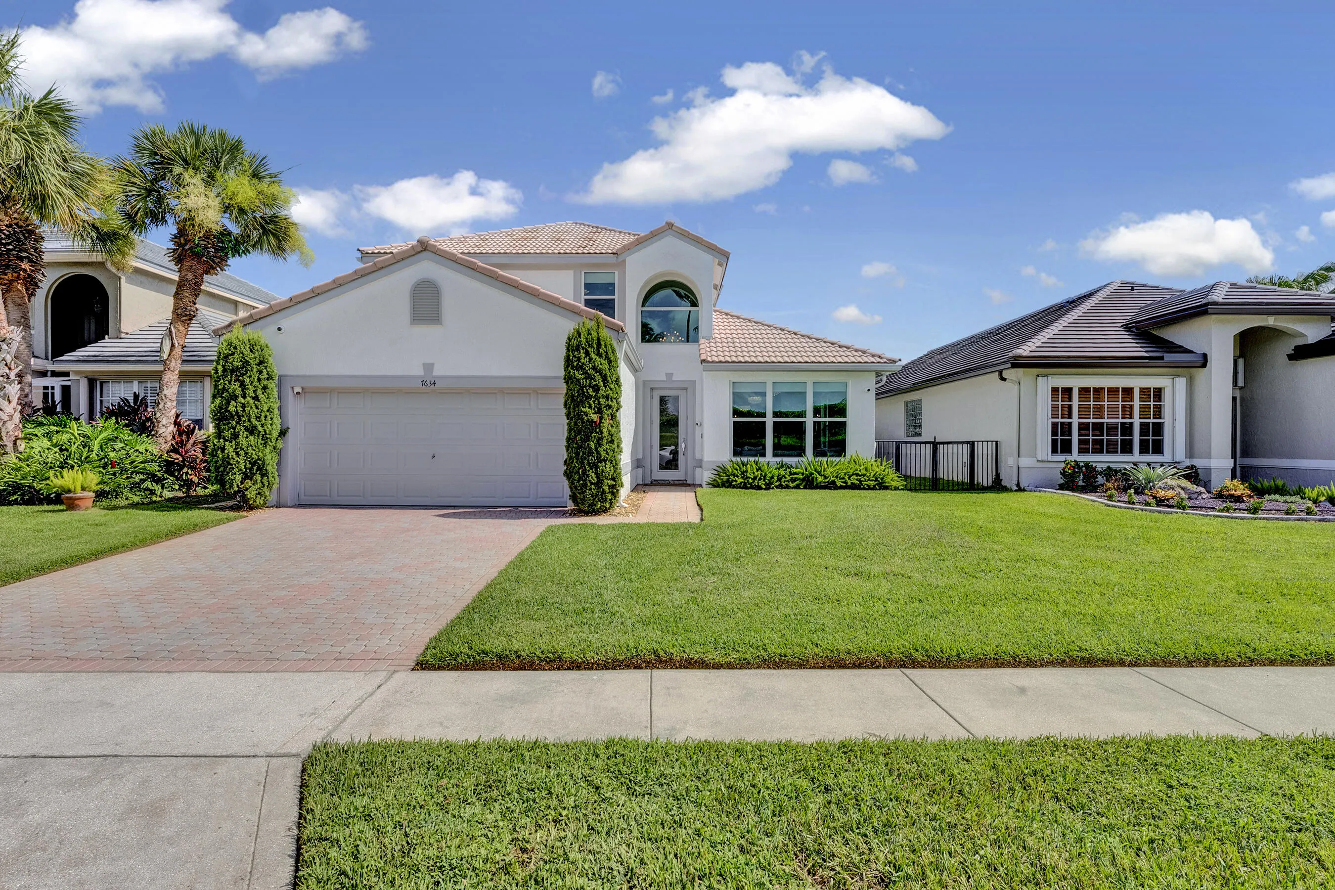 Front elevation with manicured lawn, brick paver driveway, two-car garage, and mature palm trees under blue Florida skies.