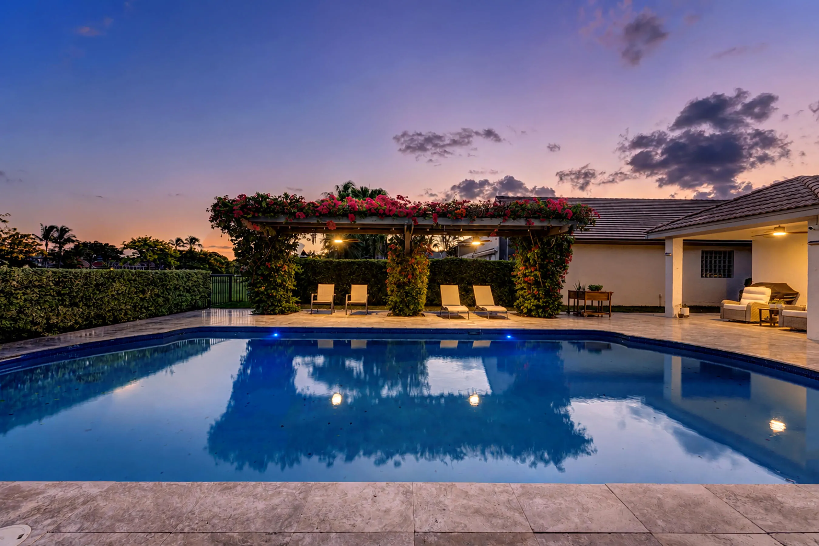 Illuminated pool and flowering pergola at dusk with lounge seating on the travertine deck.
