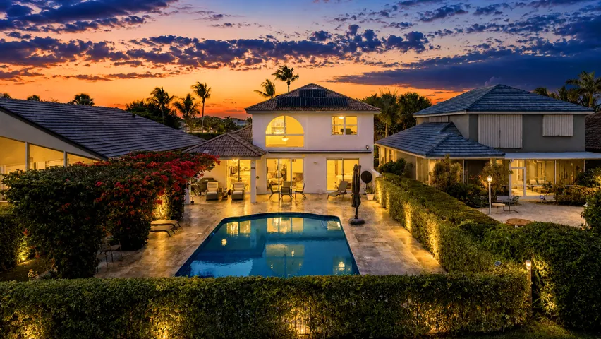 Twilight aerial view of the illuminated pool, travertine patio, and bougainvillea-covered pergola behind this Delray Beach home.