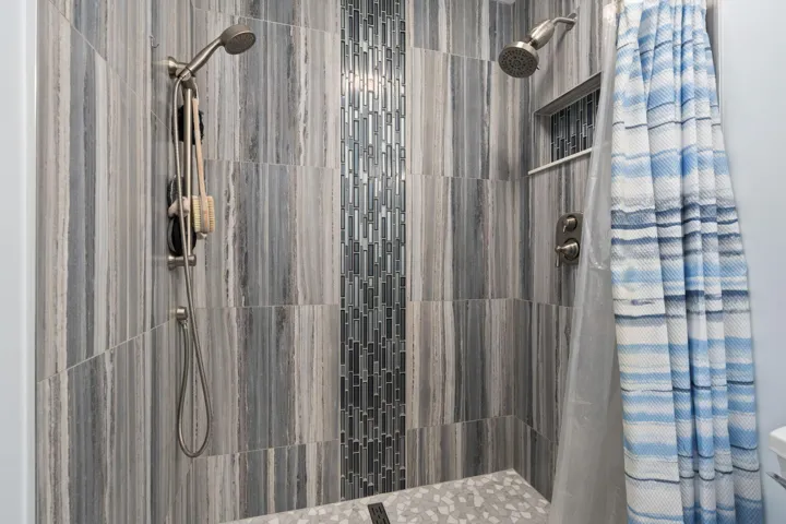 Shower enclosure featuring large format stone-look tile walls, a vertical mosaic tile accent, and dual shower heads