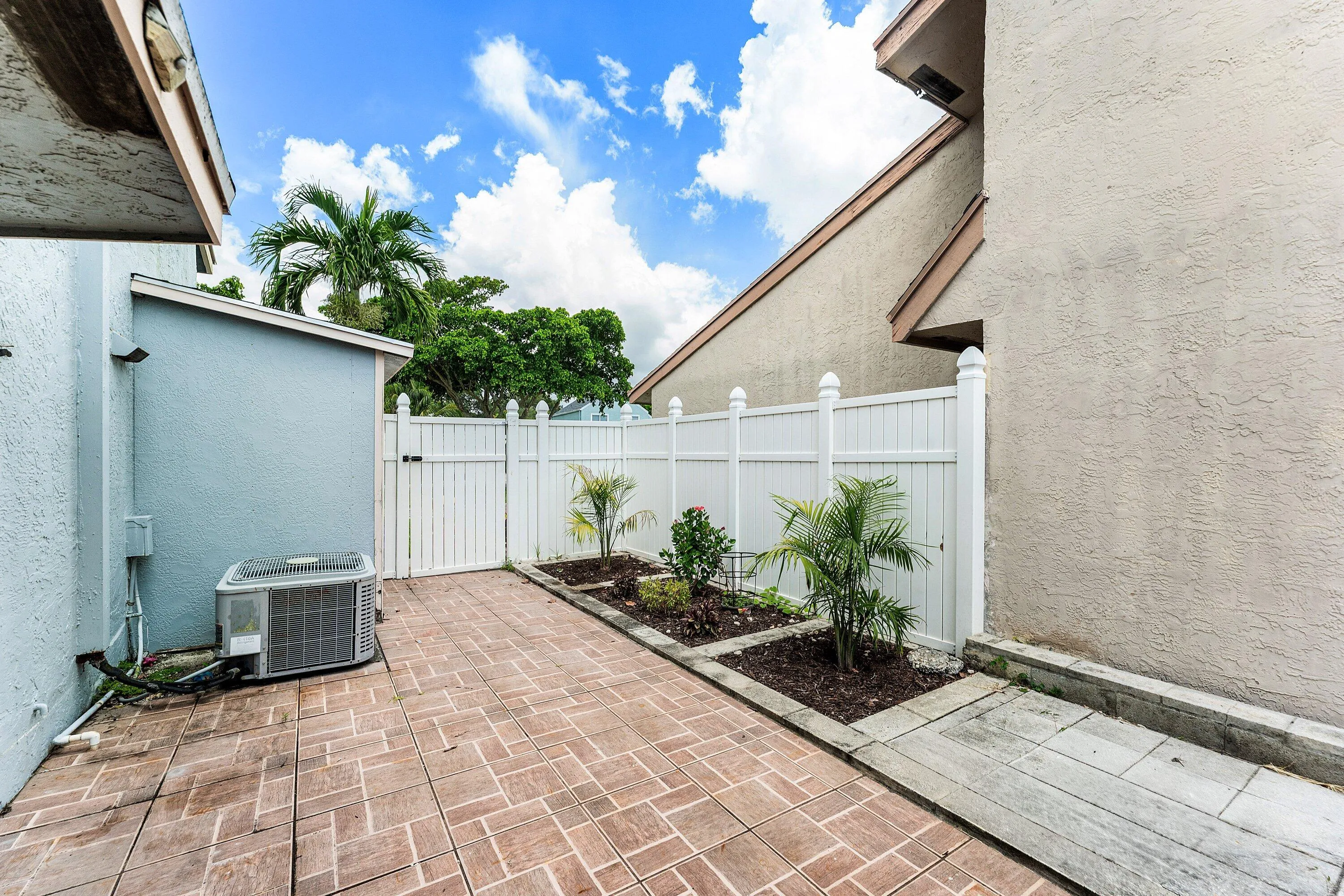 Patio area featuring brick paver flooring, white privacy fencing, and tropical landscaping