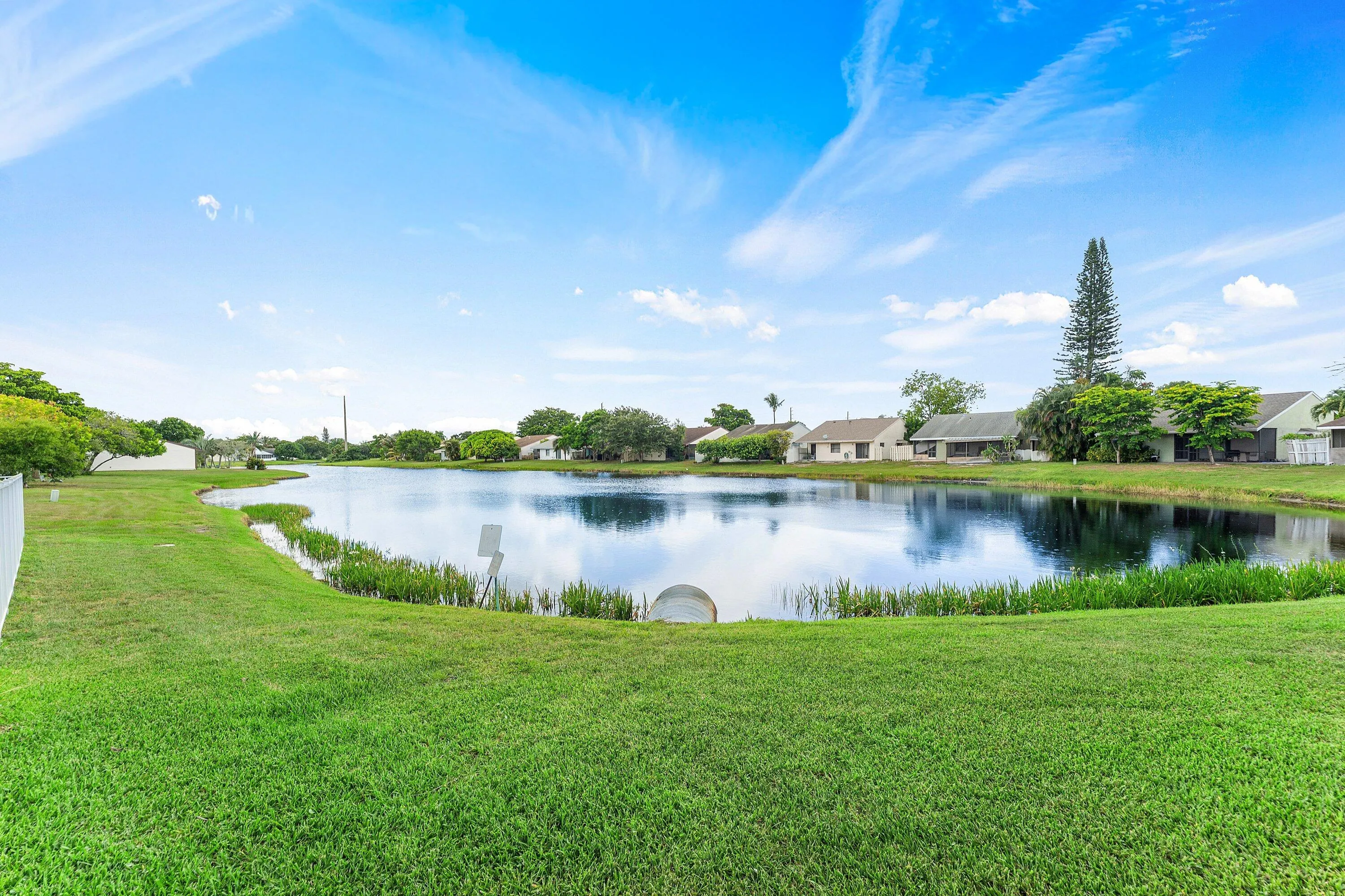 Expansive backyard featuring a large pond, lush green lawn, white perimeter fencing, and mature trees