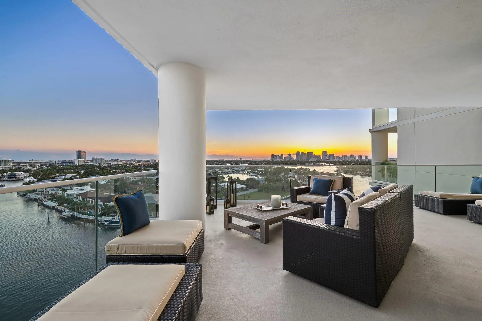 Expansive concrete balcony with glass railing, curved architectural column, and water frontage at sunset.