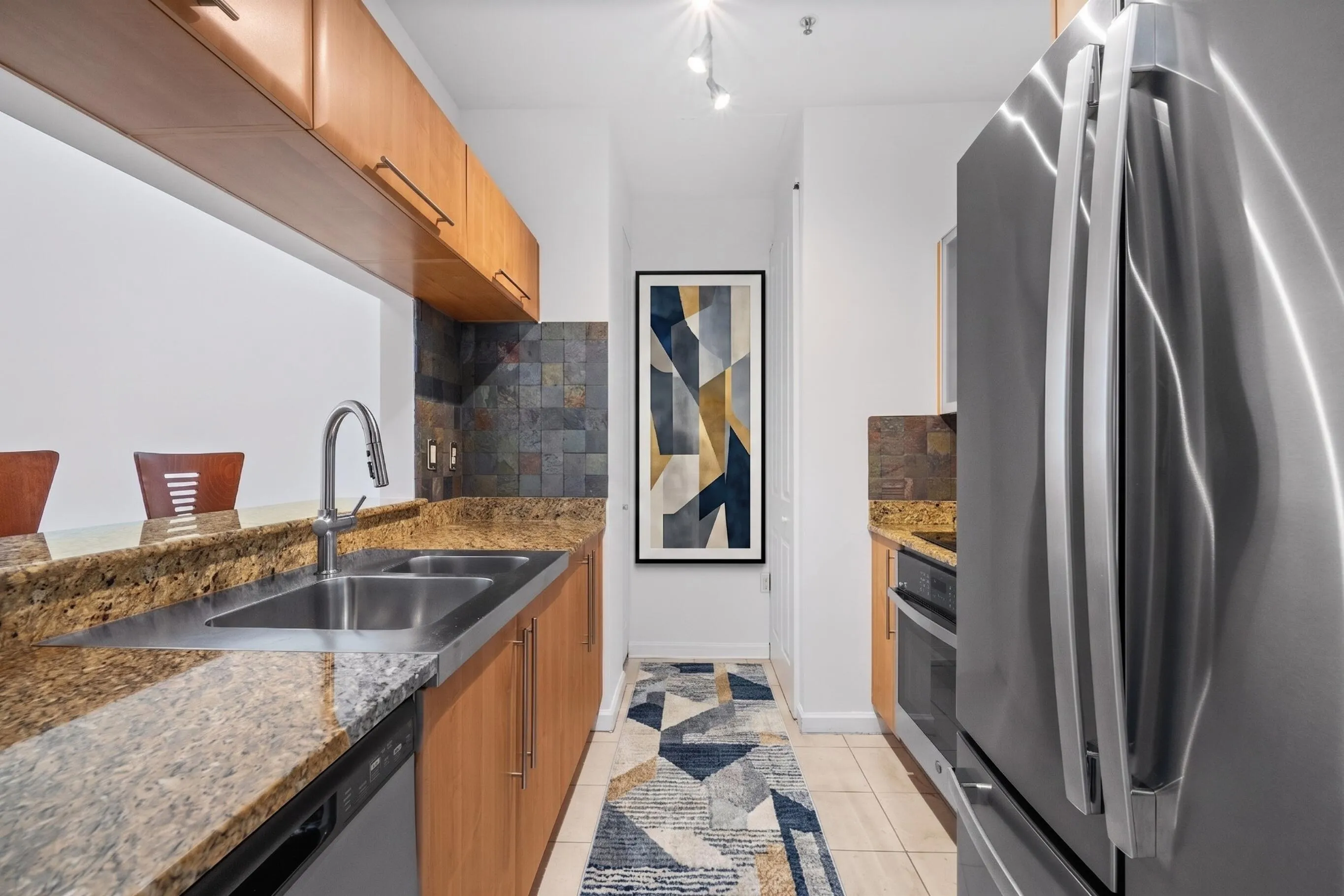 Well-appointed galley kitchen with granite countertops, warm-toned cabinetry, natural slate backsplash & newer stainless-steel appliances.