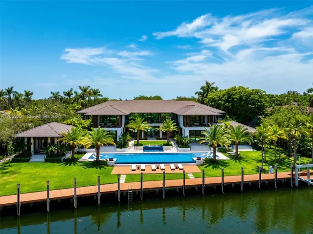 Two-story residence featuring a tile roof, an expansive rectangular pool with a built-in water feature, a sprawling lawn with mature palm trees, and a private wooden dock
