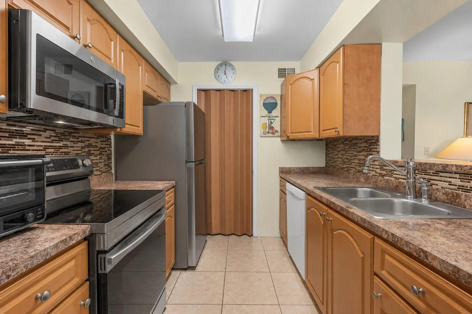 Galley-style kitchen equipped with stainless steel appliances, generous cabinetry, tile flooring, and a mosaic tile backsplash.