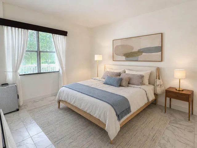 Bedroom featuring a window with blinds and sheer white curtains, light-toned walls, and large format tile flooring