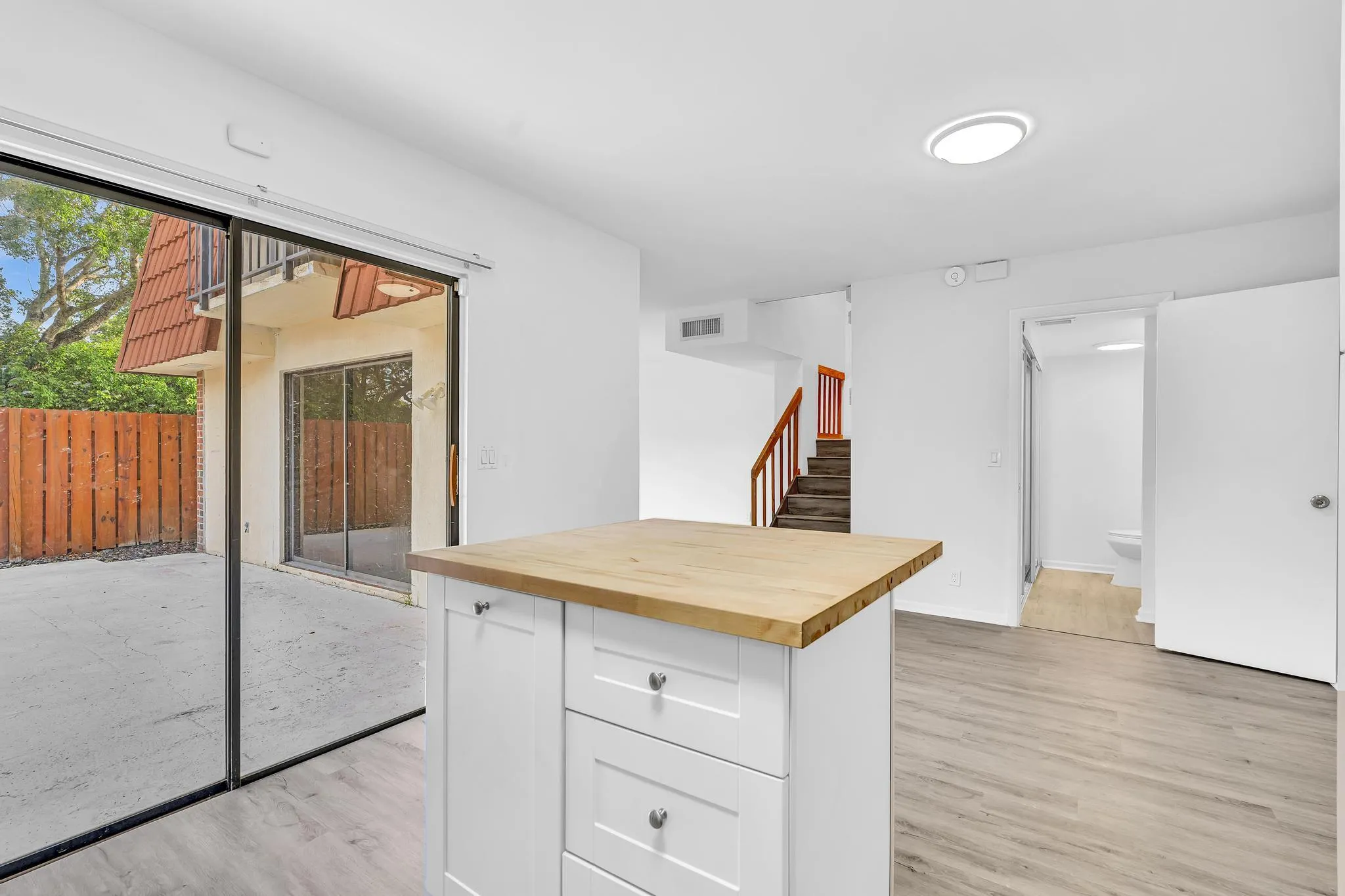 Open-concept living space featuring a central kitchen island with a wood-finish countertop, light wood-finish flooring, and a large sliding glass door providing access to an outdoor patio with a privacy fence