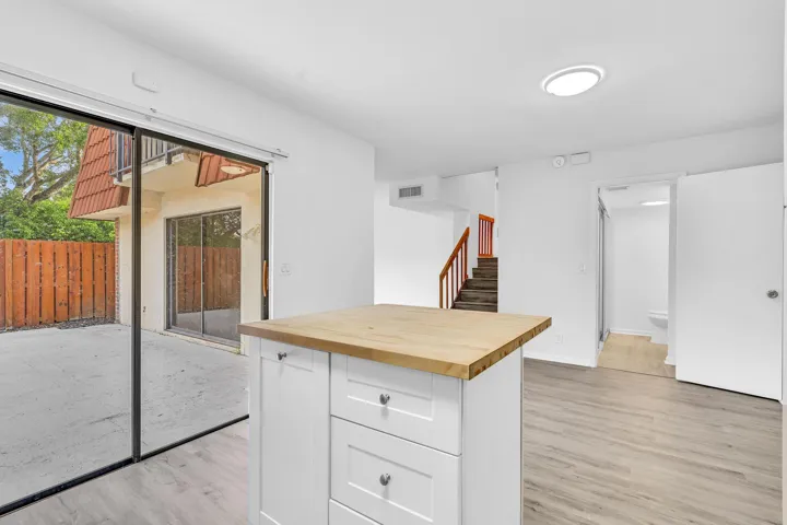 Open-concept living space featuring a central kitchen island with a wood-finish countertop, light wood-finish flooring, and a large sliding glass door providing access to an outdoor patio with a privacy fence