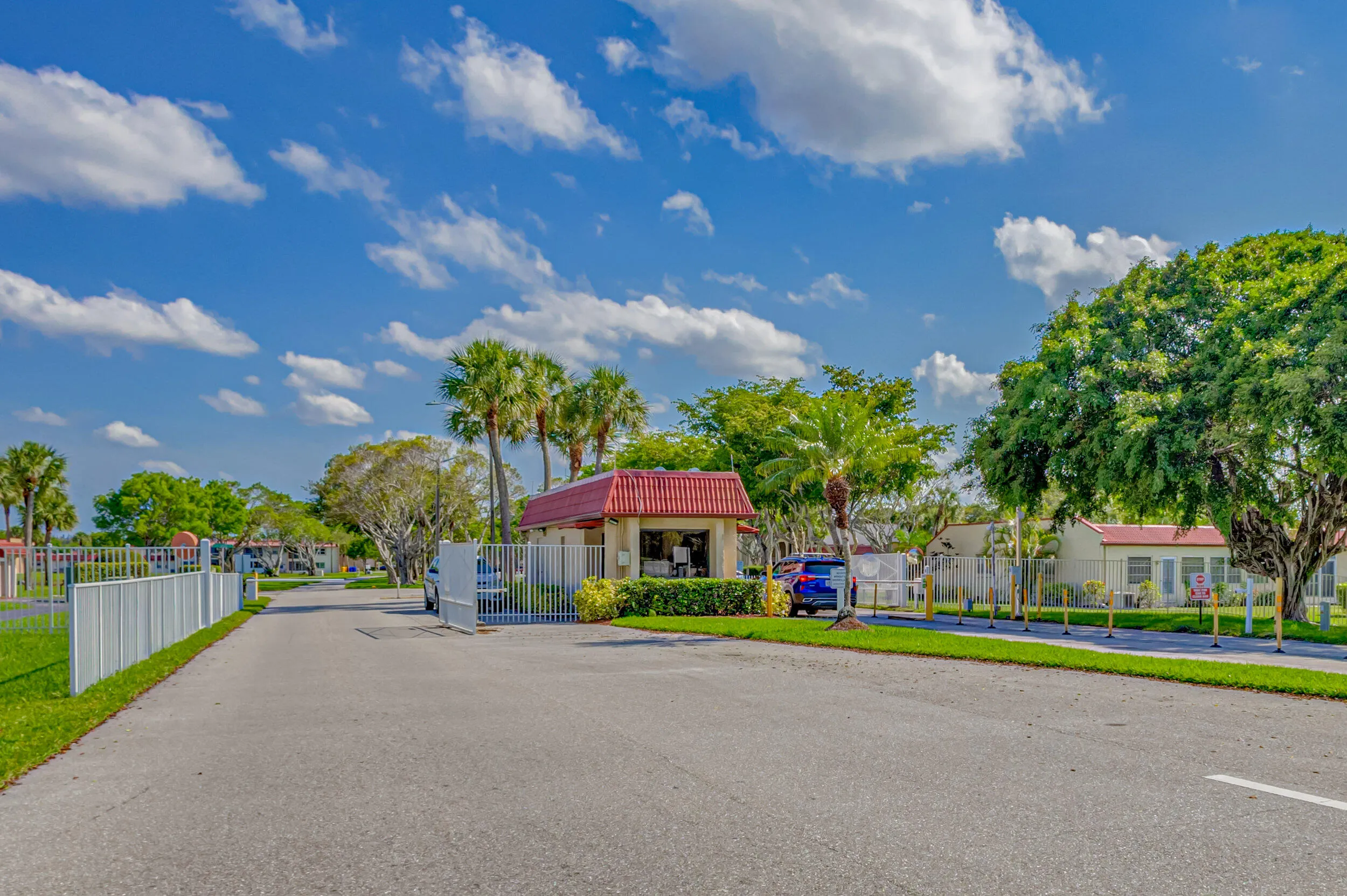 Gated community entrance featuring a security booth with a red tile roof, perimeter fencing, and mature palm trees