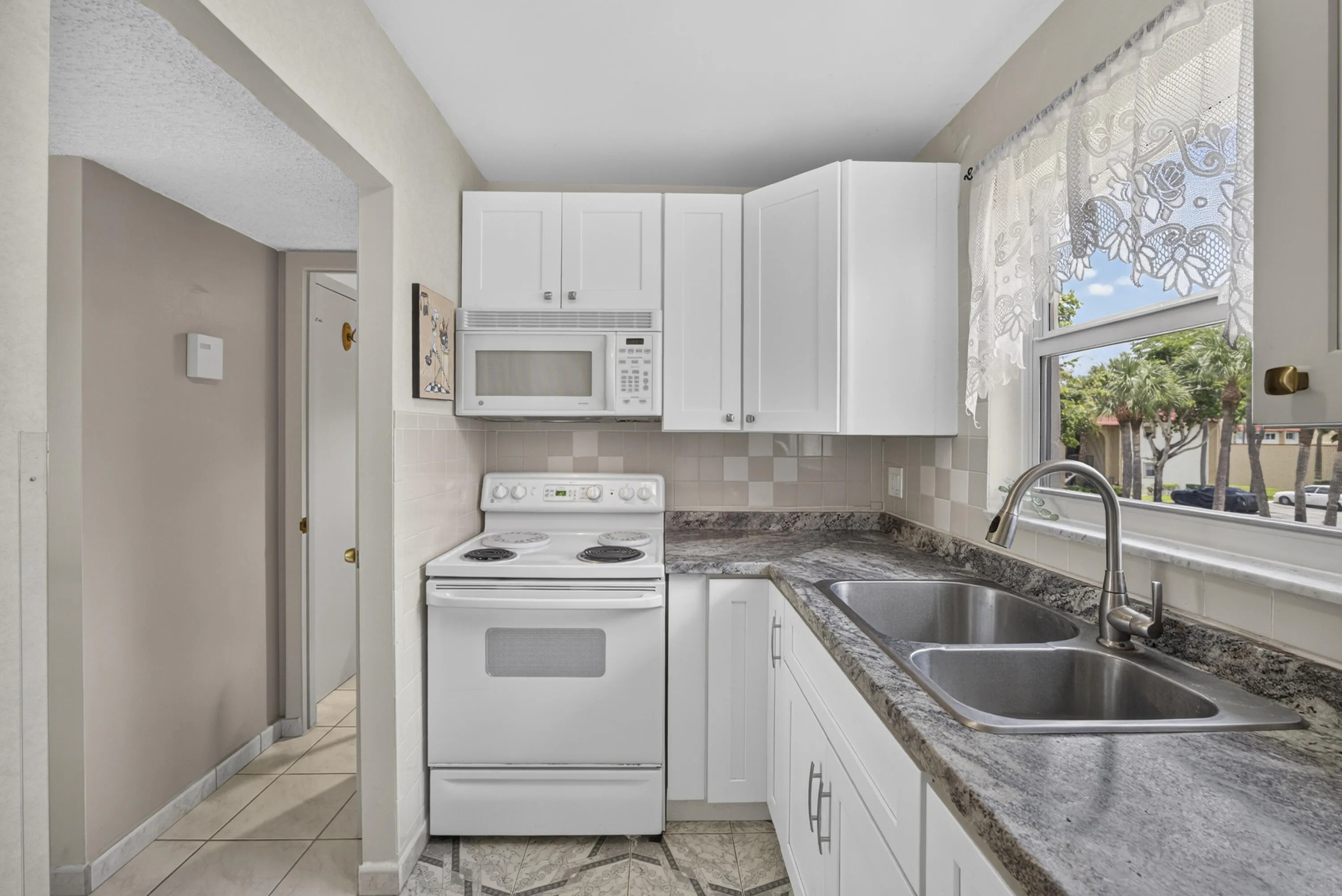 Kitchen featuring white shaker cabinetry, patterned countertop, stainless steel double basin sink with a gooseneck faucet, white electric range, and an over-the-range microwave
