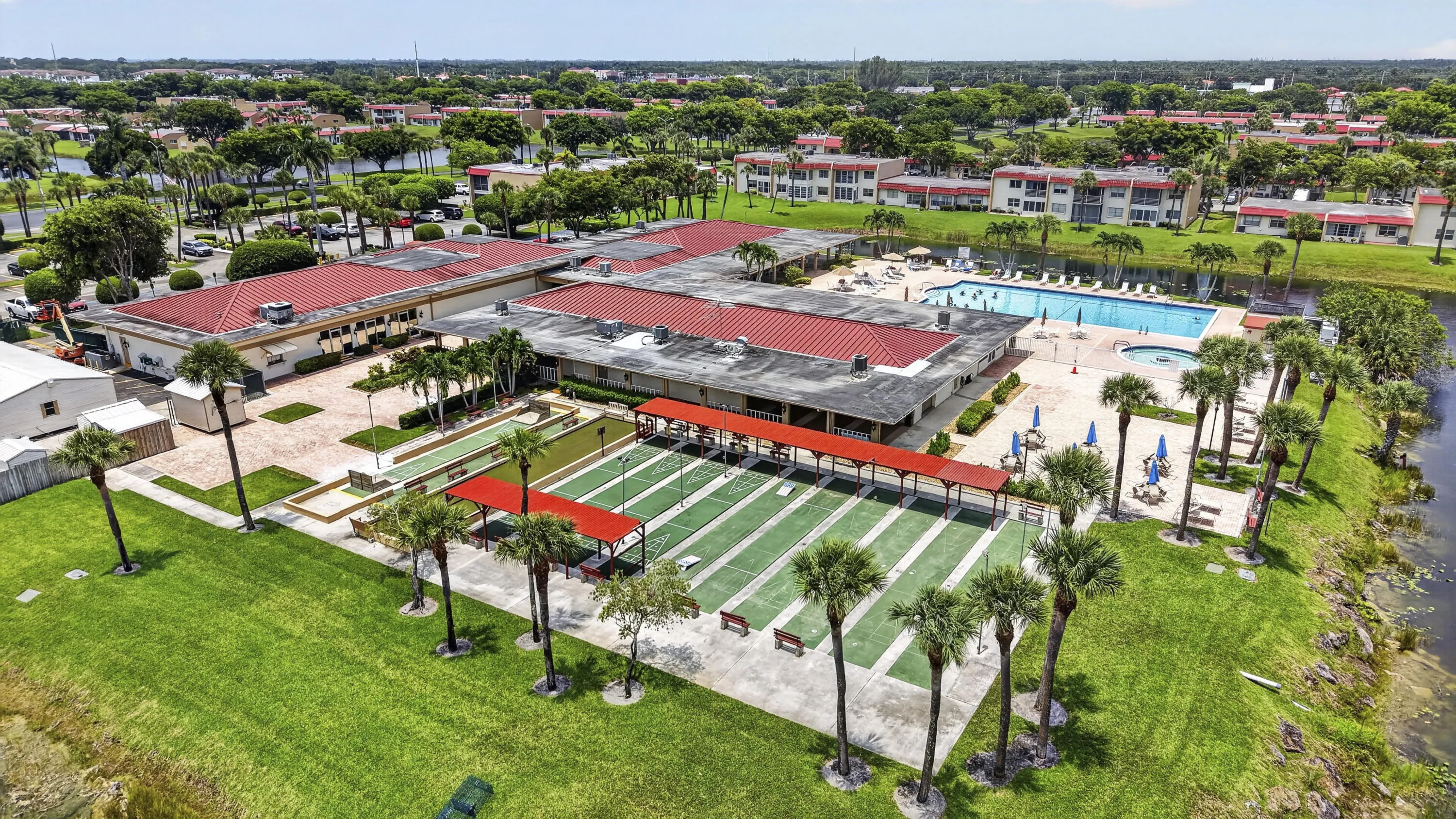 Community amenities featuring multiple buildings with red tile roofs, an outdoor swimming pool with sun deck, multiple shuffleboard courts, and mature palm trees