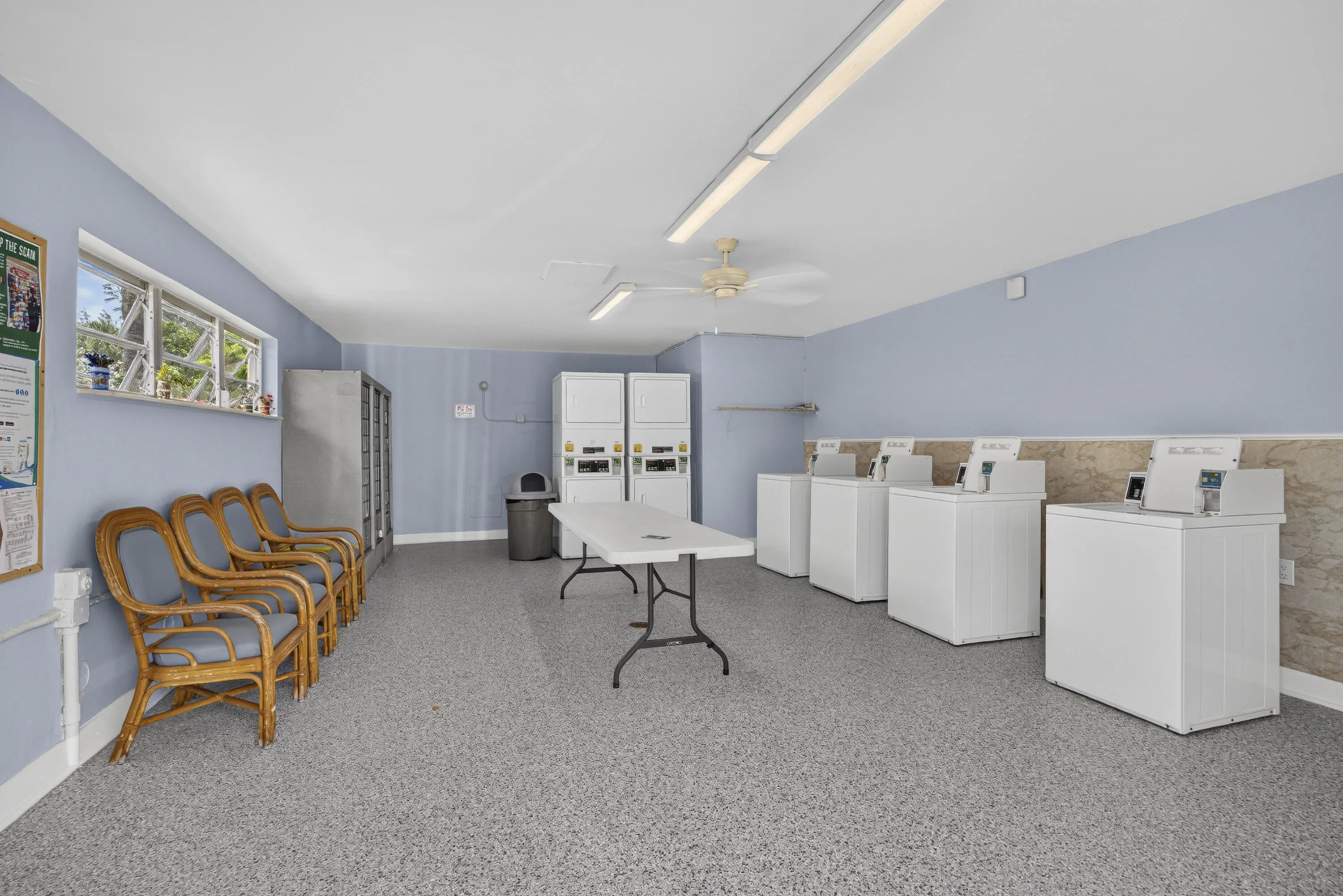 Community laundry facility featuring multiple washing machines and dryers, durable speckled flooring, and a bank of windows providing natural light