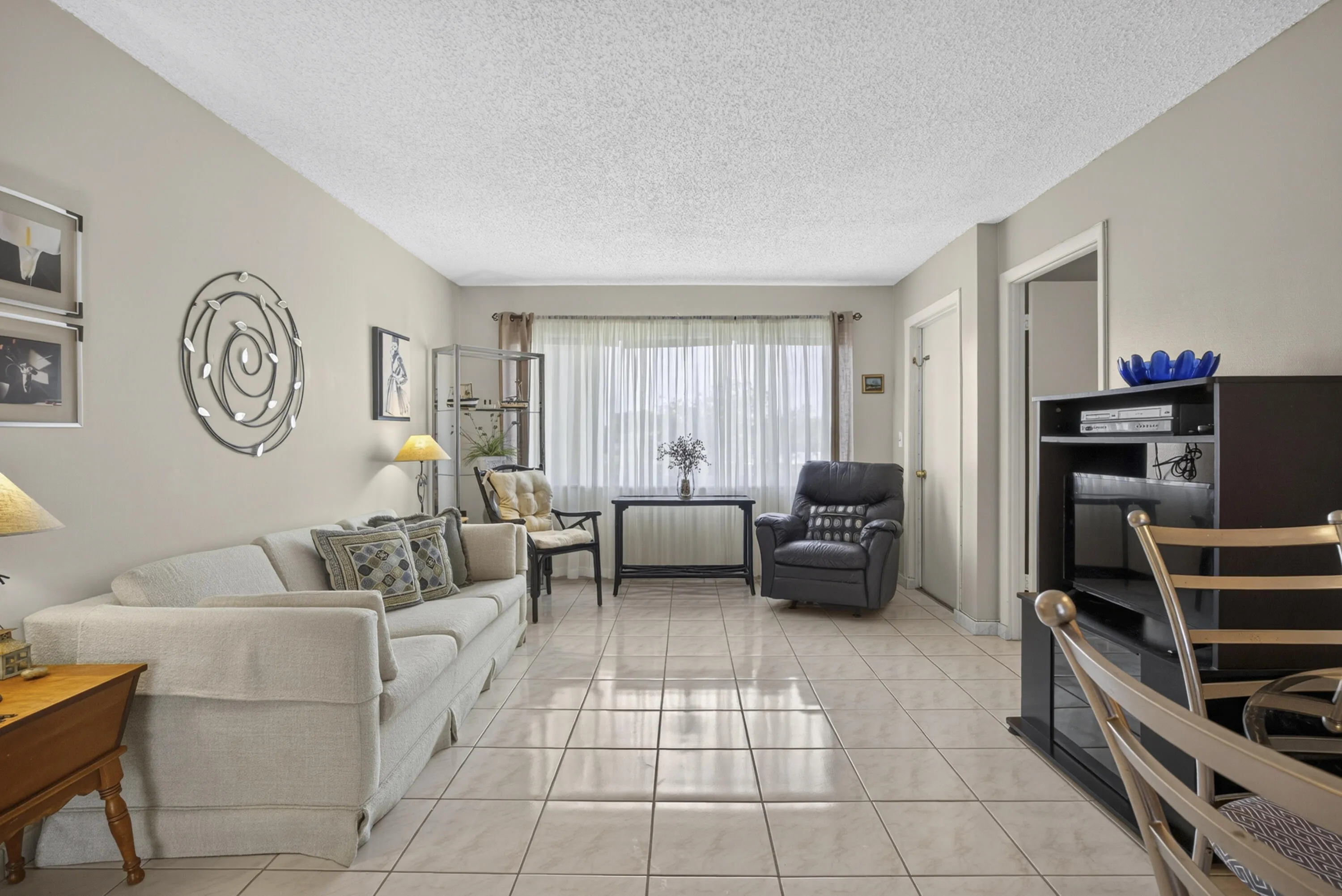 Spacious living area featuring light-toned tile flooring, neutral wall tones, and a large window with sheers