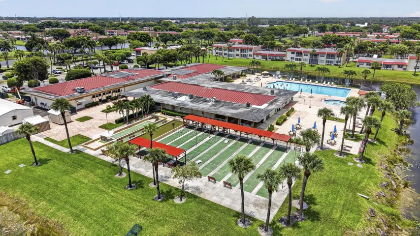 Community amenities featuring multiple buildings with red tile roofs, an outdoor swimming pool with sun deck, multiple shuffleboard courts, and mature palm trees