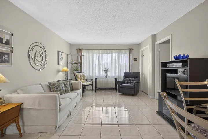 Spacious living area featuring light-toned tile flooring, neutral wall tones, and a large window with sheers