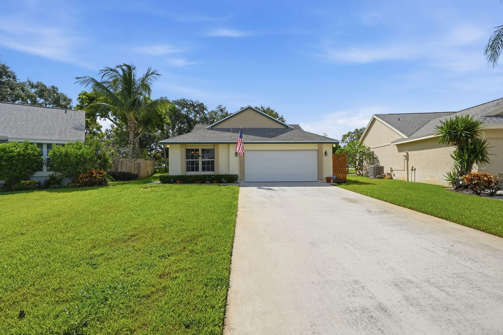 Warm beige stucco exterior with a gray shingle roof, featuring a two-car garage, a concrete driveway, and a verdant lawn