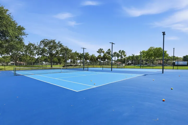 Blue tennis court featuring white line markings, a net, and surrounded by green landscaping with mature trees and palm trees