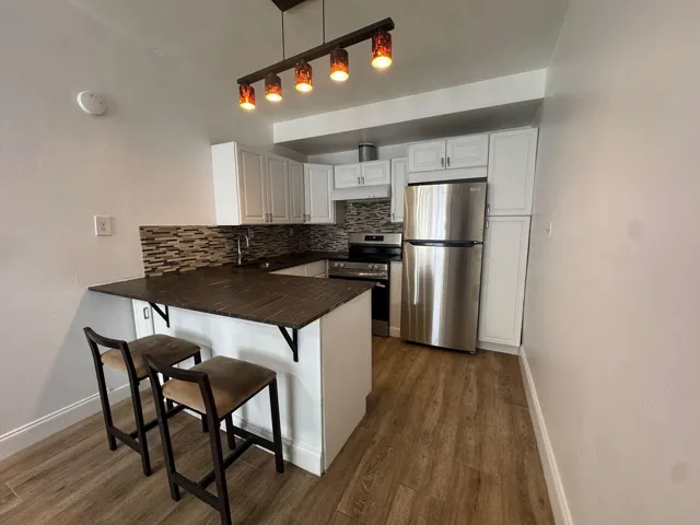 Contemporary kitchen featuring white cabinetry, stainless steel appliances, a mosaic tile backsplash, wood-finish flooring, and a breakfast bar with dark wood countertop