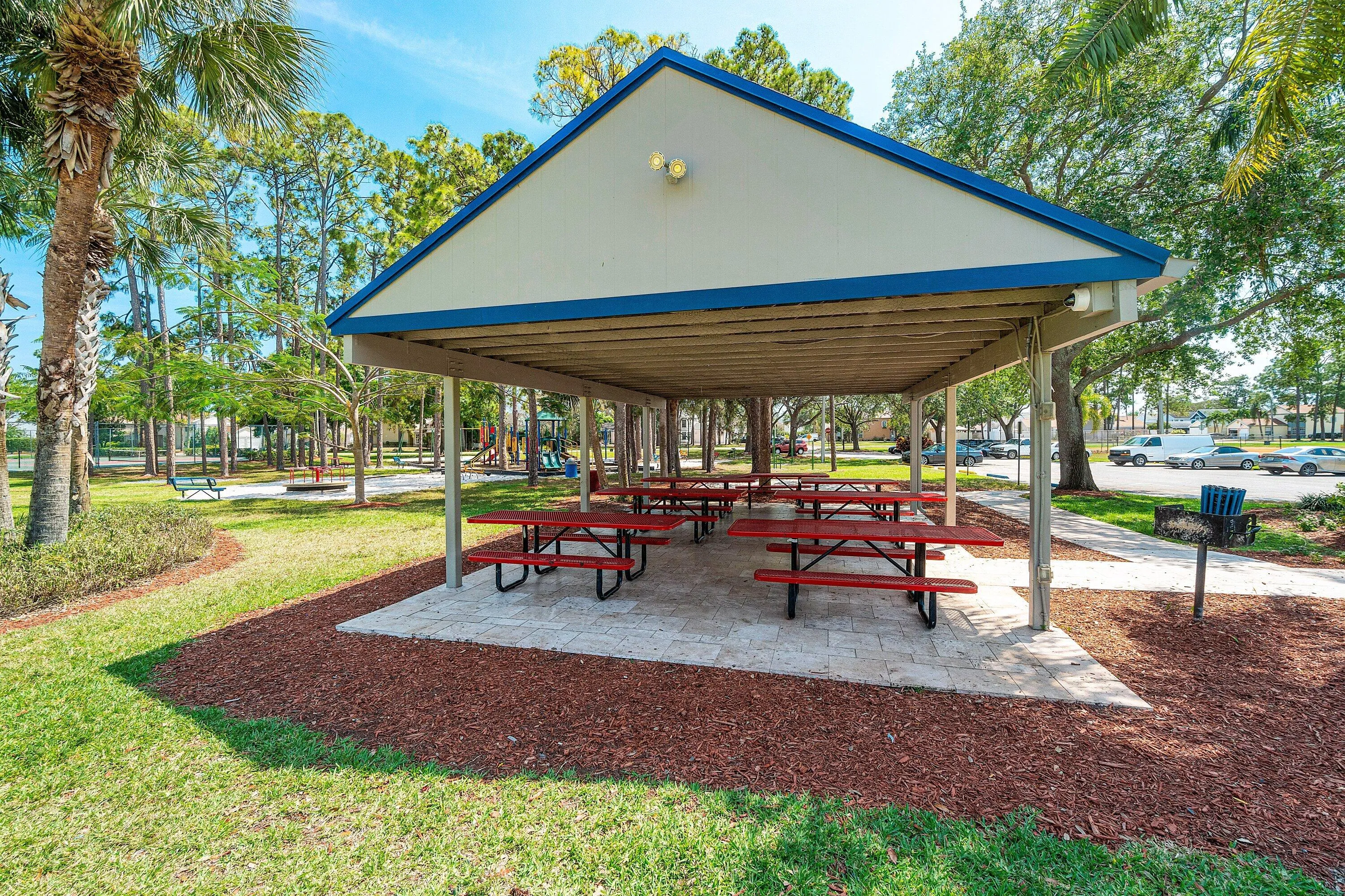 Community pavilion featuring a light-colored roof with blue trim, multiple red picnic tables, a built-in grill, and surrounding green lawn with palm trees