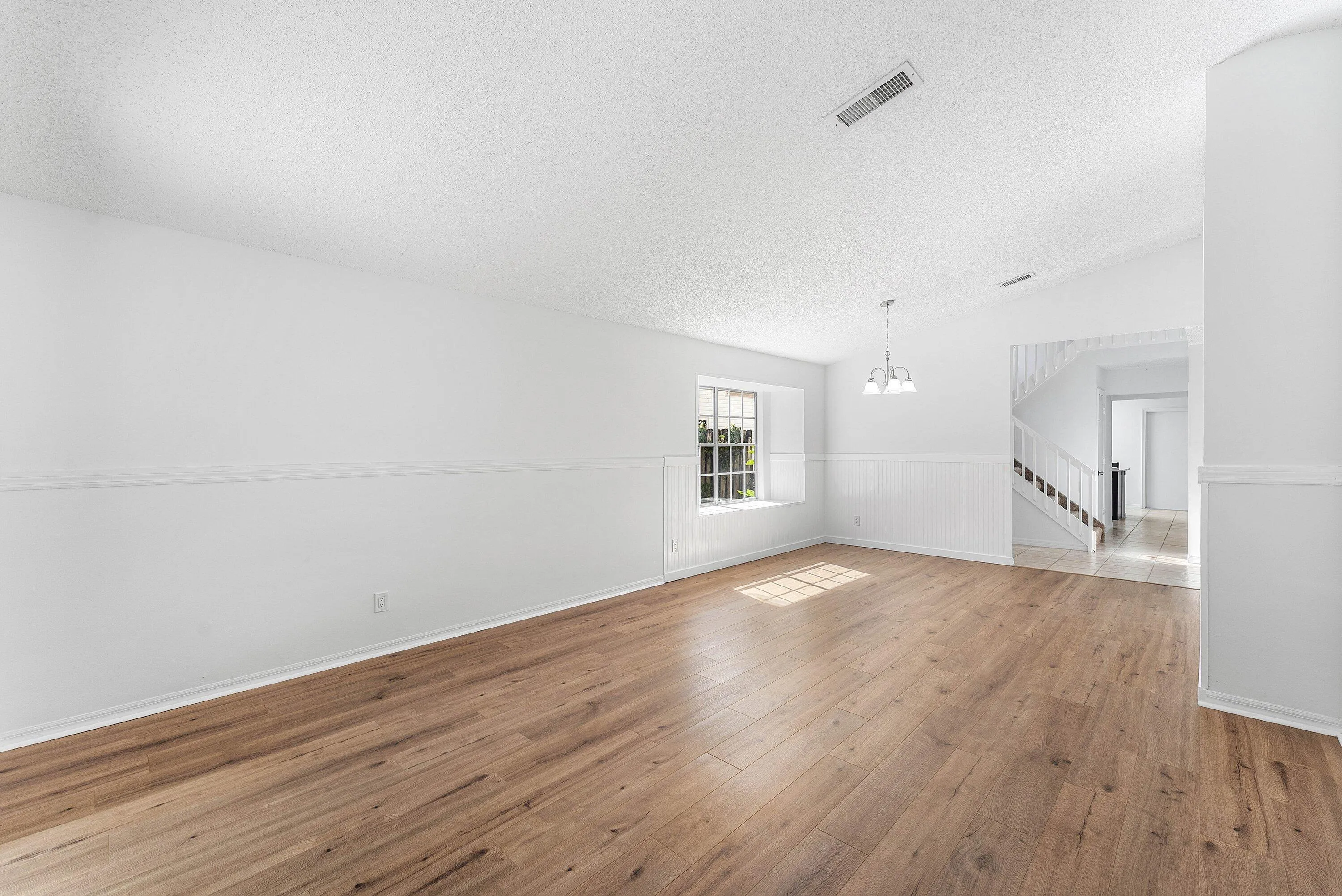 Spacious room featuring wood-finish flooring, white walls with wainscoting, an overhead light fixture, and a window with natural light