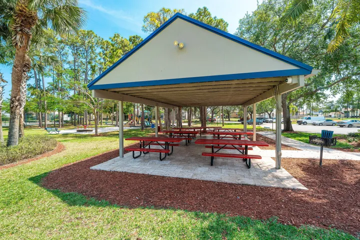 Community pavilion featuring a light-colored roof with blue trim, multiple red picnic tables, a built-in grill, and surrounding green lawn with palm trees