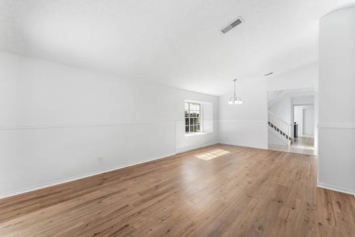 Spacious room featuring wood-finish flooring, white walls with wainscoting, an overhead light fixture, and a window with natural light