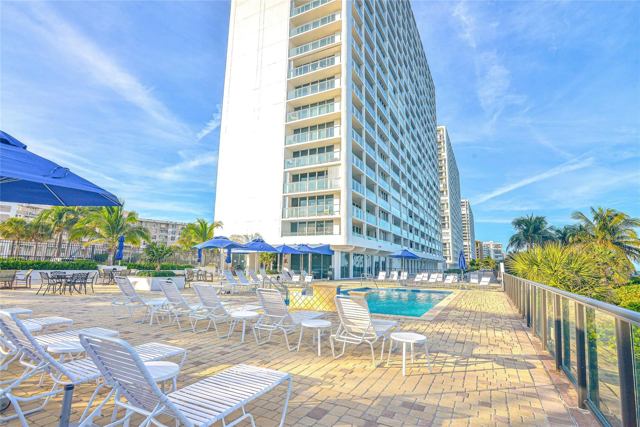 Outdoor amenity deck featuring a swimming pool, paved sundeck, multiple seating areas with umbrellas, and a glass-paneled railing system