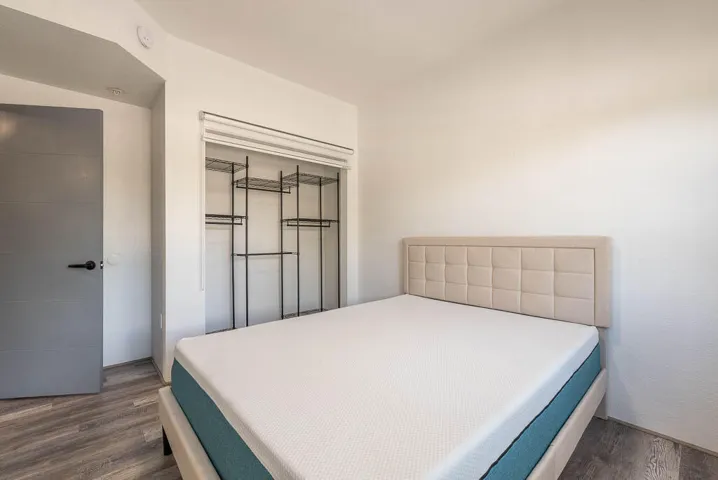 Sleeping area featuring light wood-finish flooring, a contemporary headboard, a built-in closet system with metal shelving and hanging rods, white walls, and a gray door with black hardware