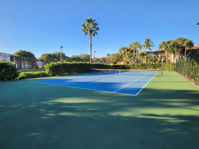Blue and green hard tennis court with white line markings, surrounded by a dark perimeter fence and mature landscaping