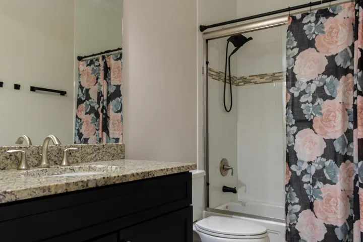 Bathroom featuring a black vanity with a light-toned granite countertop, brushed nickel sink fixtures, a full-width mirror, and a shower-over-tub with white tile surround and a decorative tile accent strip